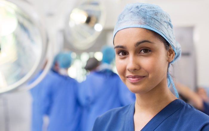 Female nurse is prominent at the front of the image as the mai focus. She is wearing blue hospital scrubs and a cap. The background has nurses (out of focus) operating in a hospital theatre.